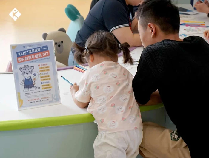 KLIS teacher crouching to guide child painting hand‑fan during community activity