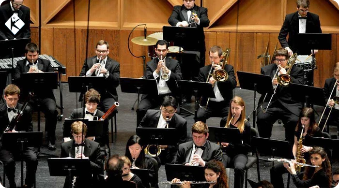 University wind ensemble performing on concert hall stage with full orchestra